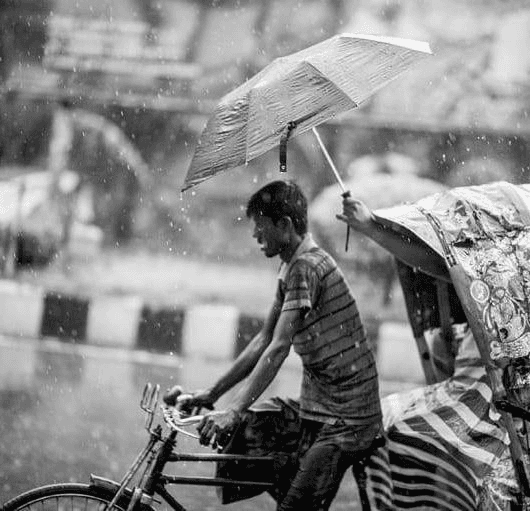 A cyclist in the rain, an umbrella held over him by the person riding behind
